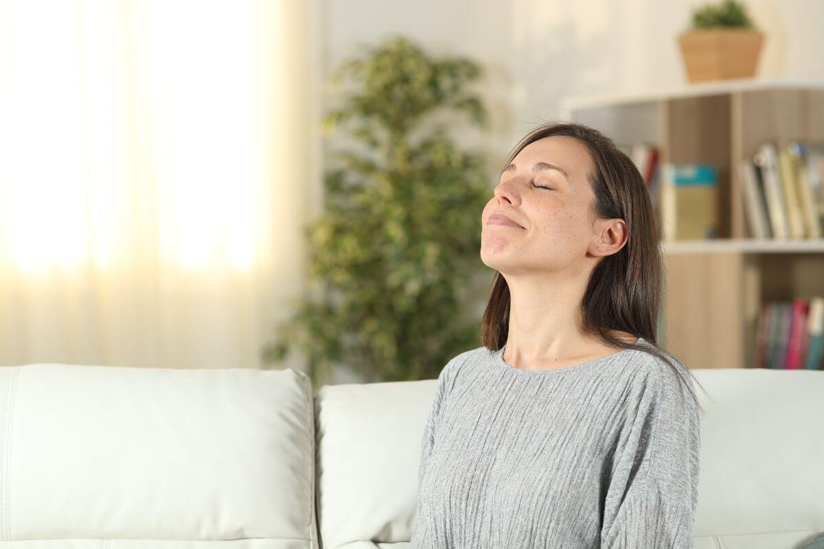 A woman with long brown hair and wearing a gray sweater sits on a white sofa with her eyes closed and a serene expression on her face.