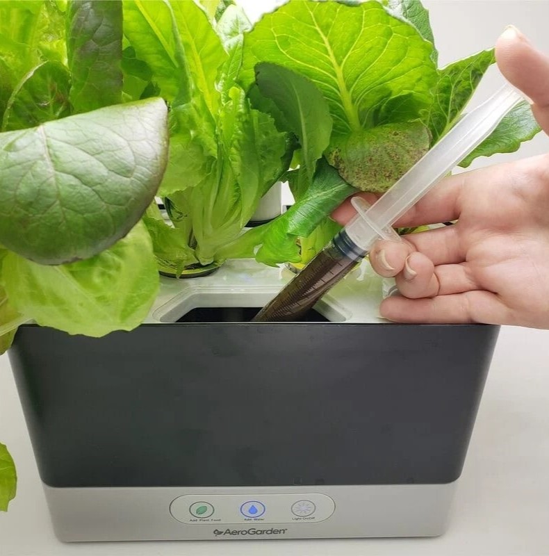 A person is injecting a brown liquid into a small indoor hydroponic garden with a large syringe. 