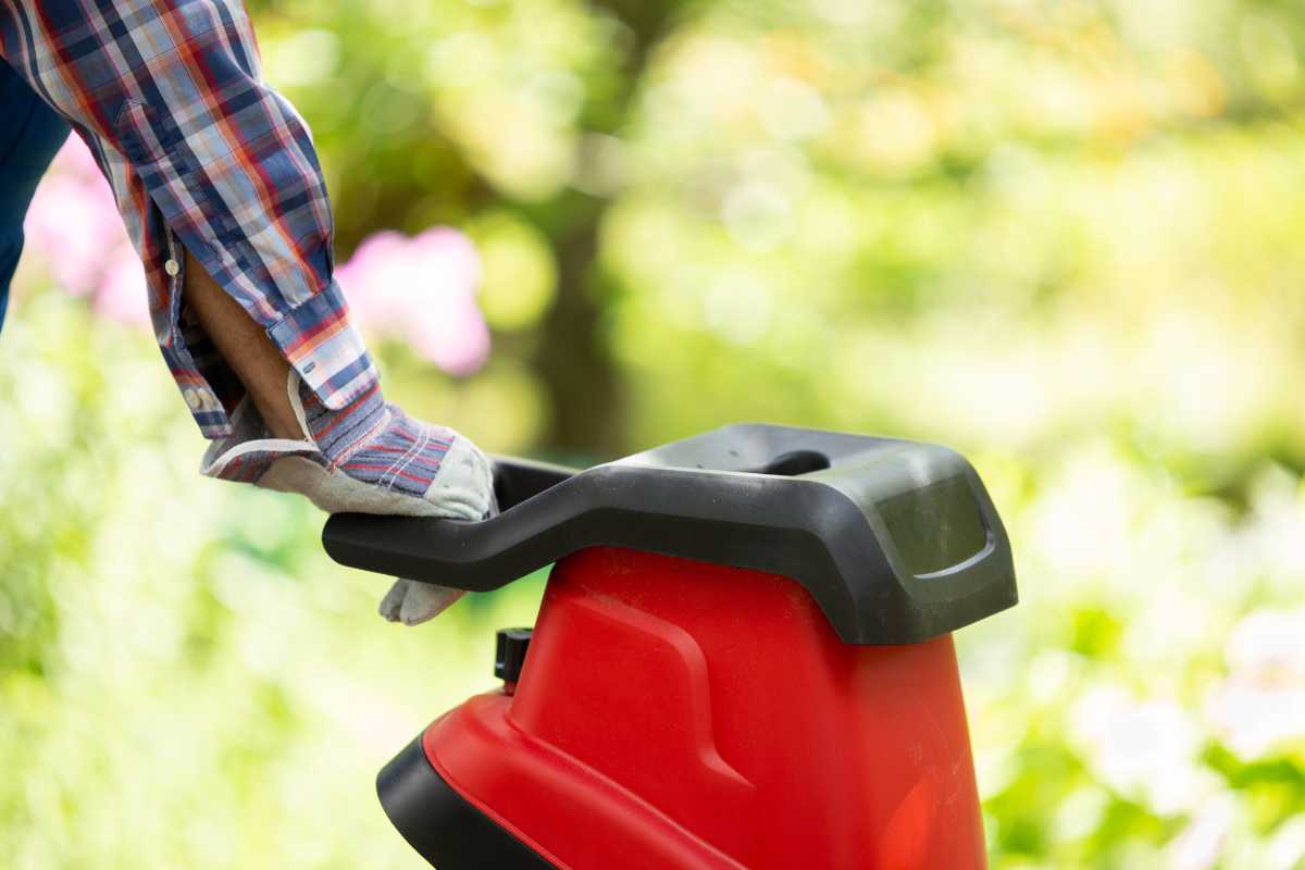 A person wearing a checkered shirt and gloves is pushing the handle of a red woodchipper.