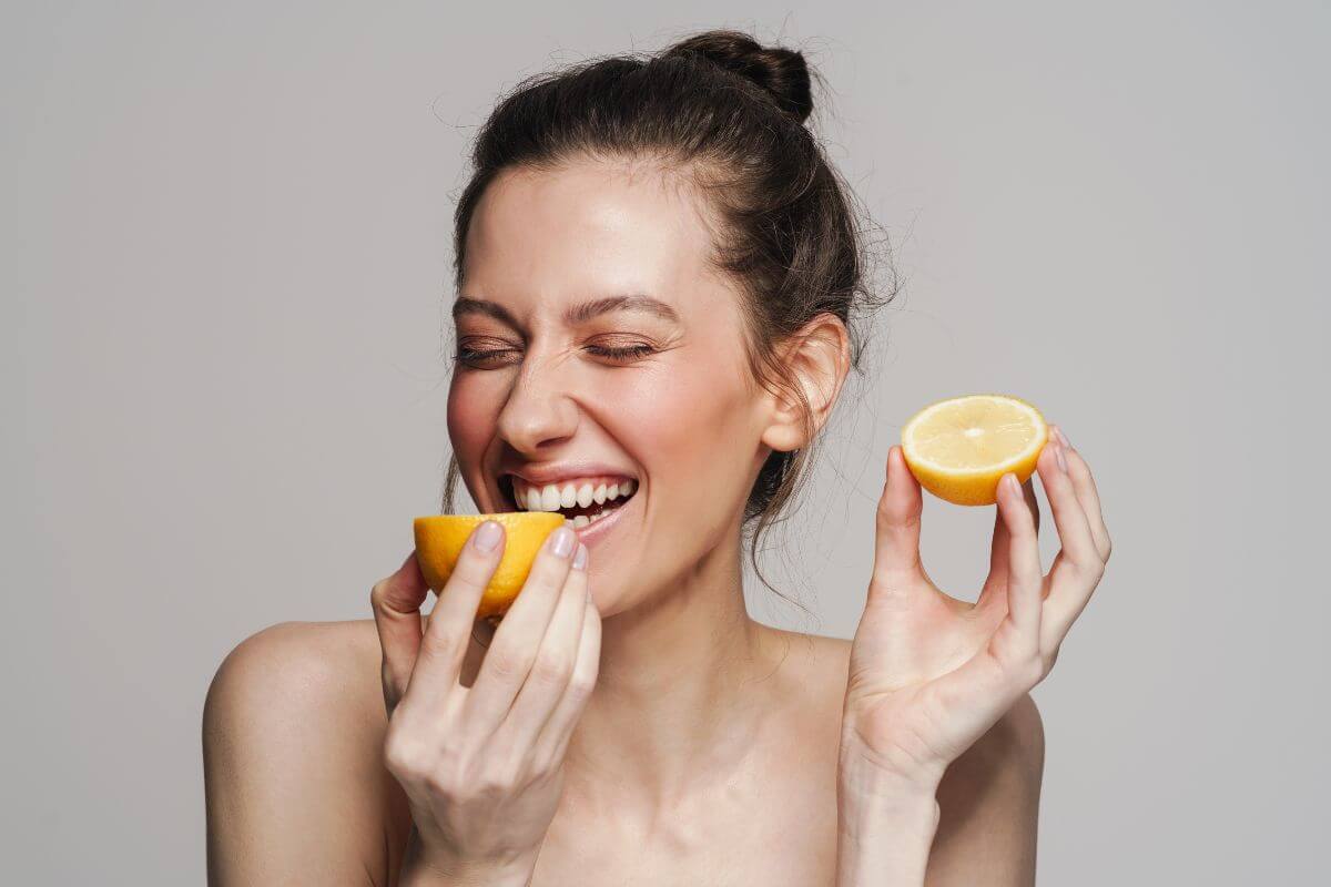 A woman with her hair in a bun holds two halves of a lemon close to her face.