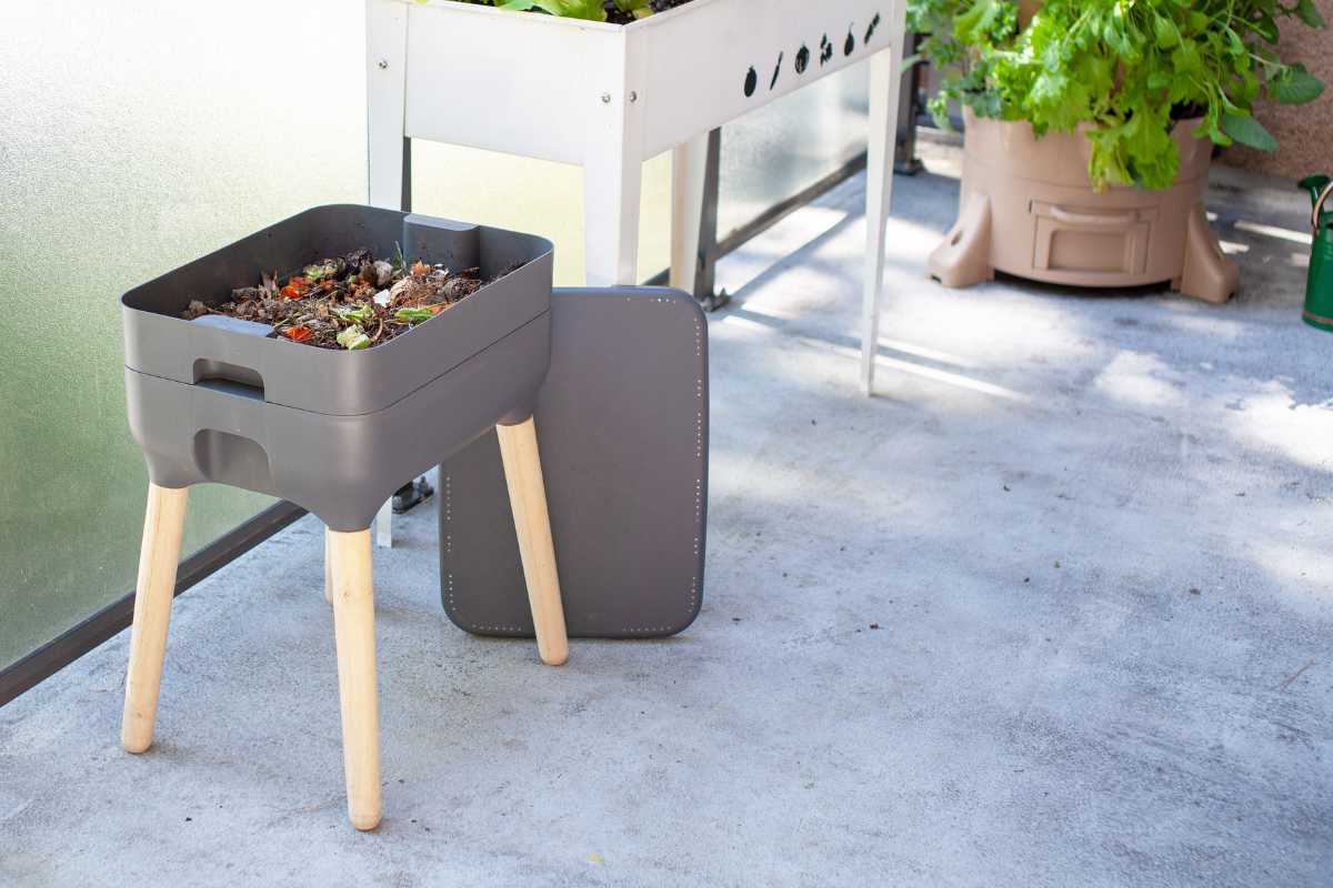 A gray, two-tiered worm composting bin with wooden legs stands on a gray patio. 