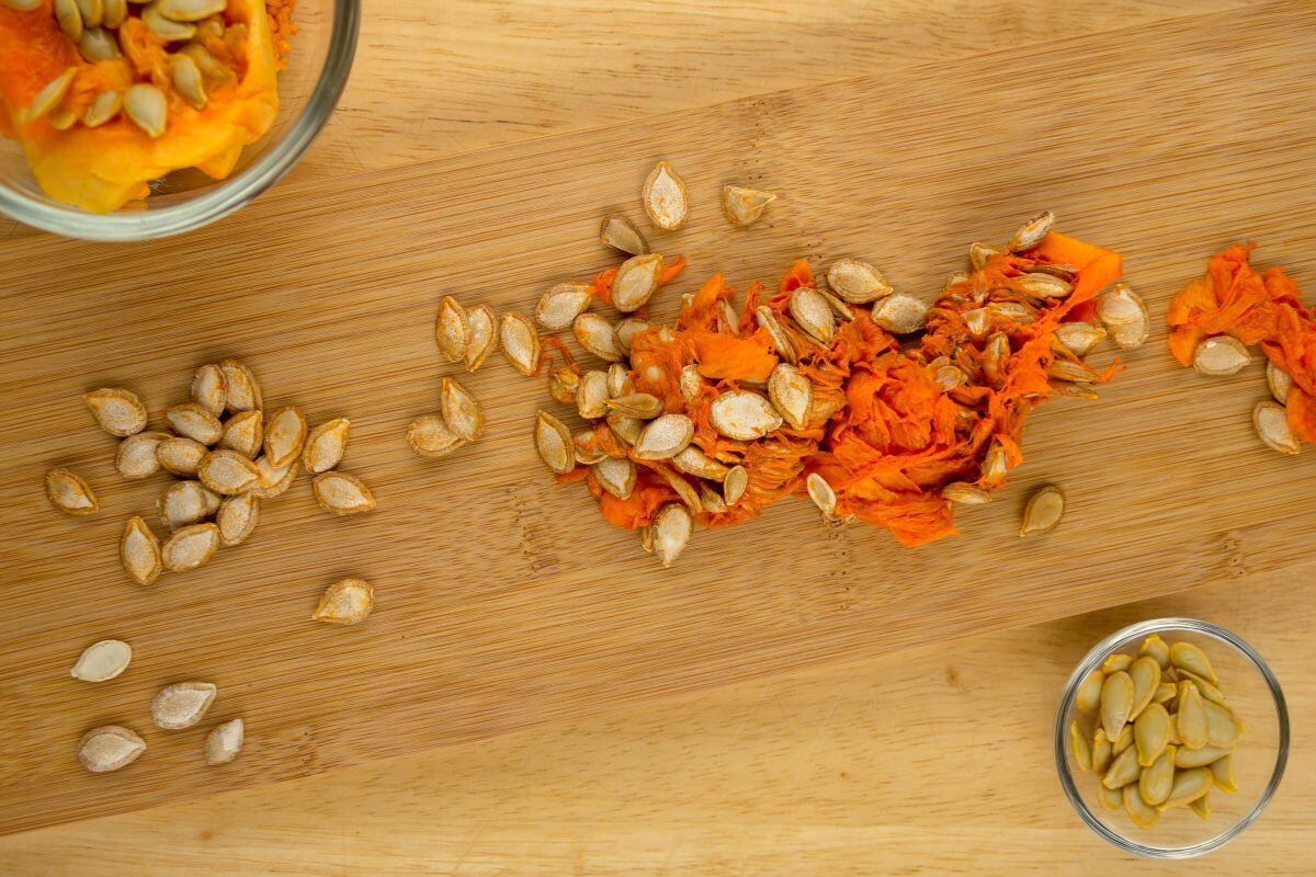 A wooden cutting board with separated squash seeds and squash flesh. Piles of seeds are on the board, alongside bright orange flesh. 