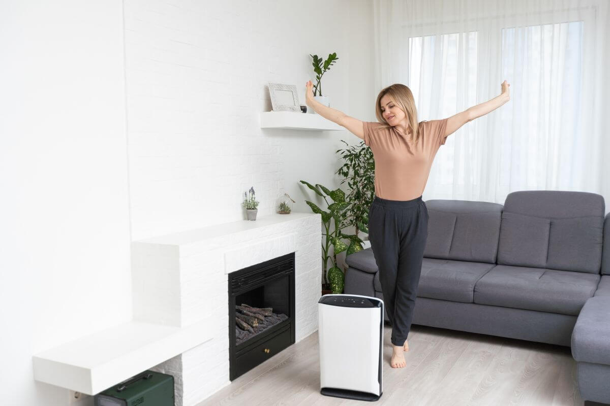 A woman stretches her arms up while standing in a modern living room with a gray sofa, white walls, and green plants.