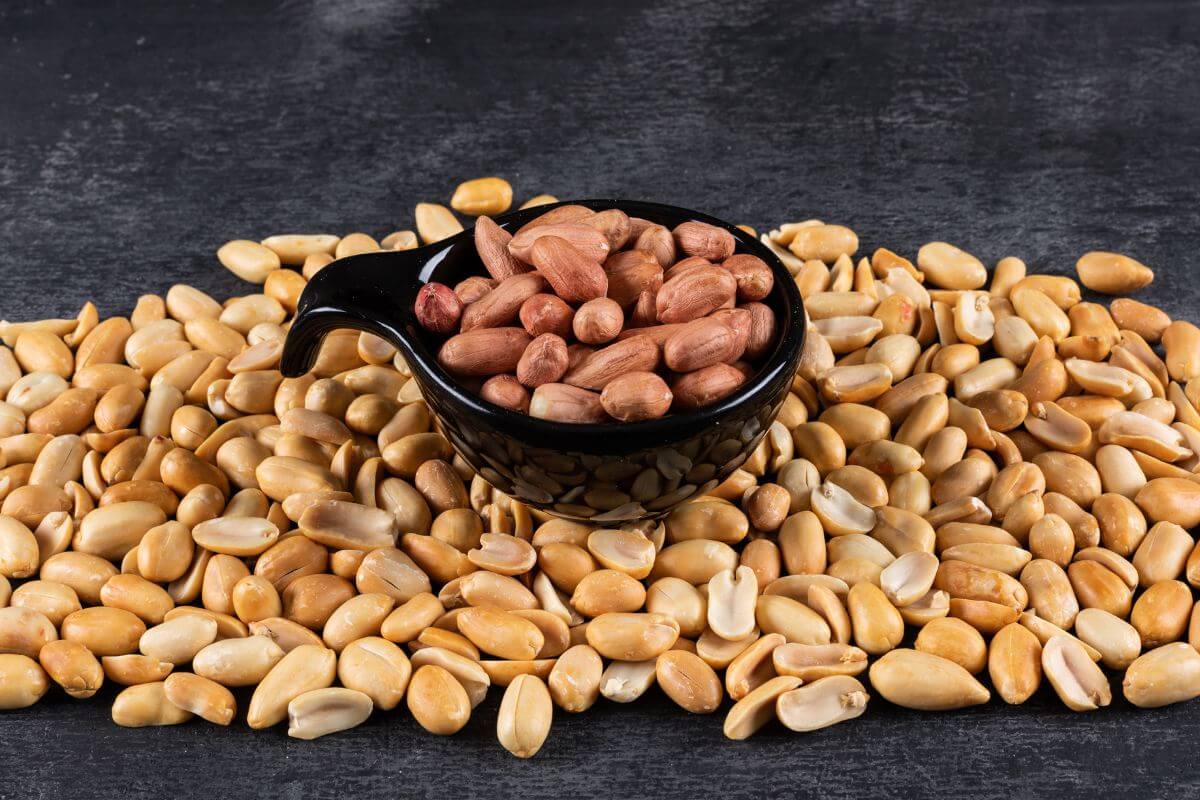 A small black cup of unshelled peanuts sits on a large pile of unshelled peanuts against a dark background.