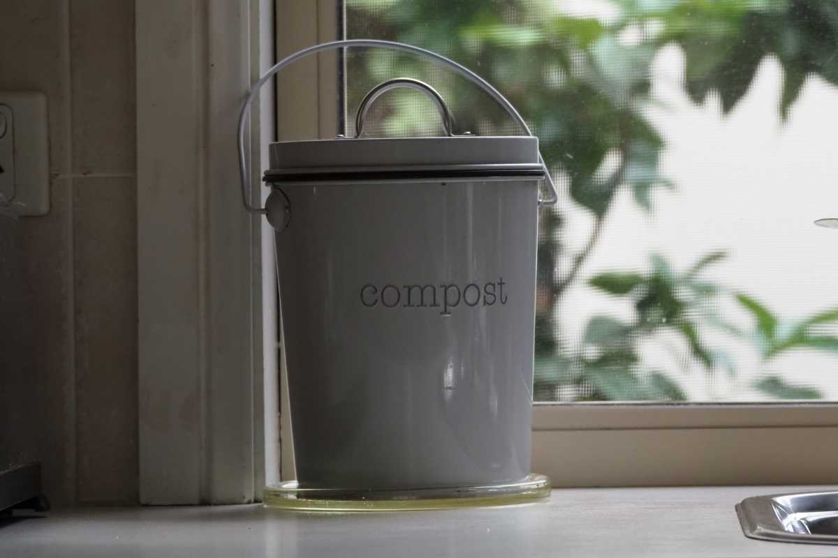 A gray metal compost bin with a handle sits on a kitchen countertop near a window. 