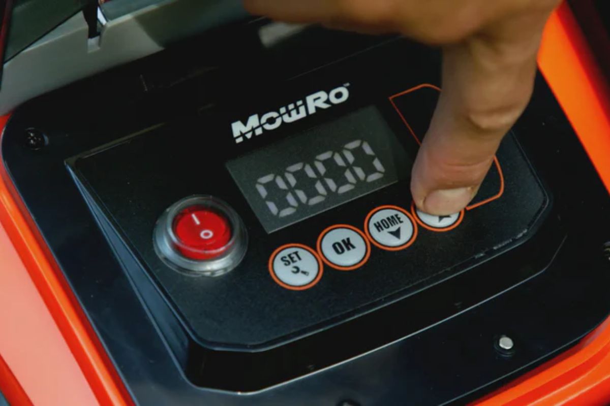 A person's finger pressing the "Home" button on a MowRo RM24 robotic lawn mower's control panel. The digital display shows "00:00," and other buttons labeled "SET," "OK," and "START" are visible.