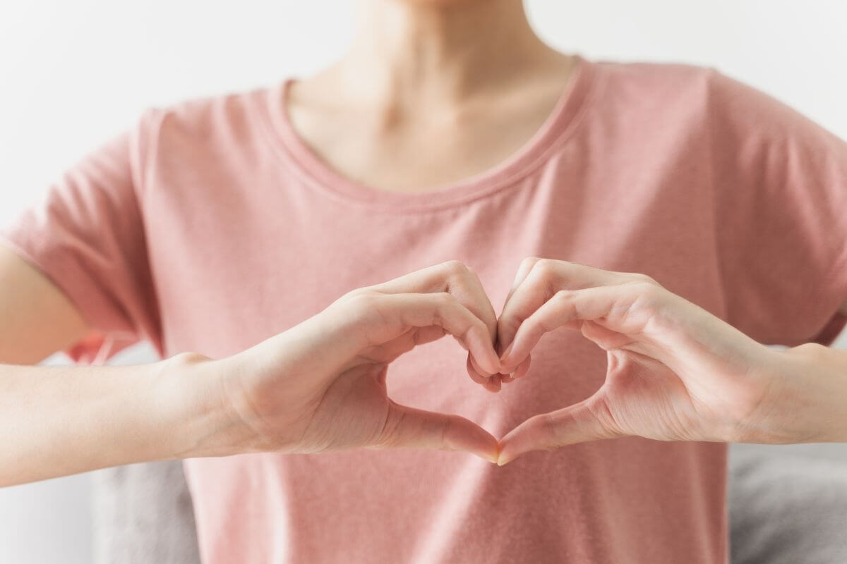 A person wearing a pink shirt is forming a heart shape with their hands in front of their chest.