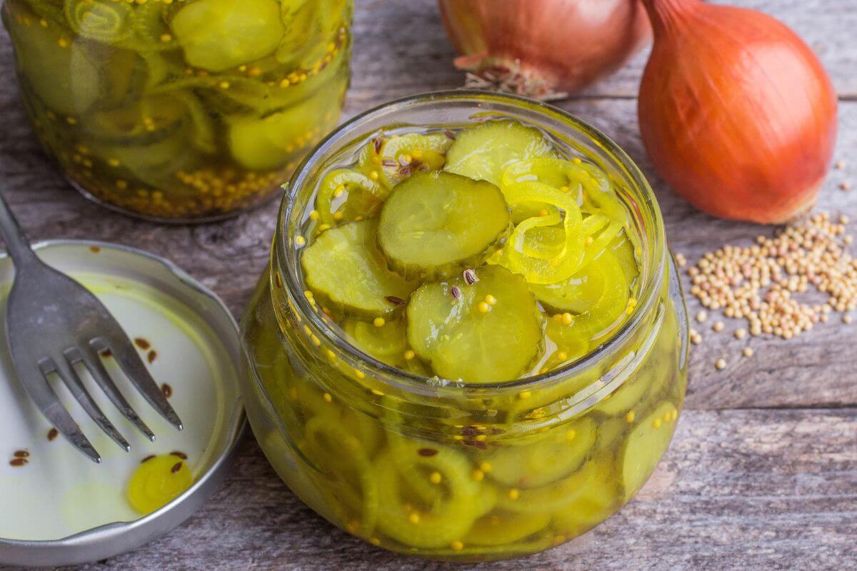 A glass jar filled with pickle slices and mustard seeds, placed on a rustic wooden surface.