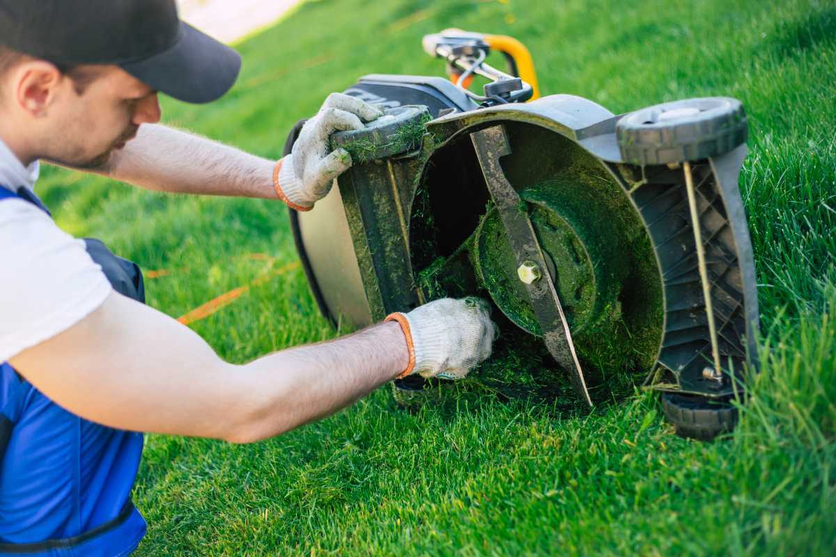 A person in a cap and gloves is performing maintenance on an overturned lawnmower on a grassy lawn. 