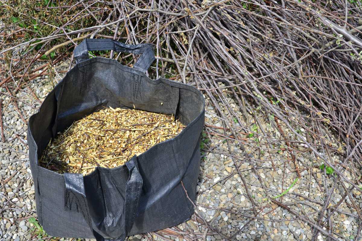 A black, reusable bag filled with wood chips is placed on a gravelly surface. In the background, there is a pile of dry branches.