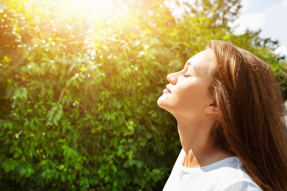 A woman with long brown hair stands outdoors with her eyes closed, basking in the sunlight and inhaling clean air. 