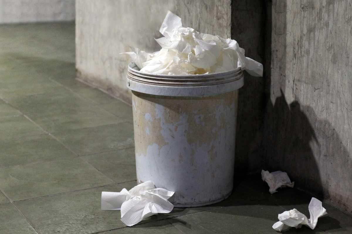 A white trash can filled with crumpled tissues and compost paper towels is placed against a rough concrete wall.