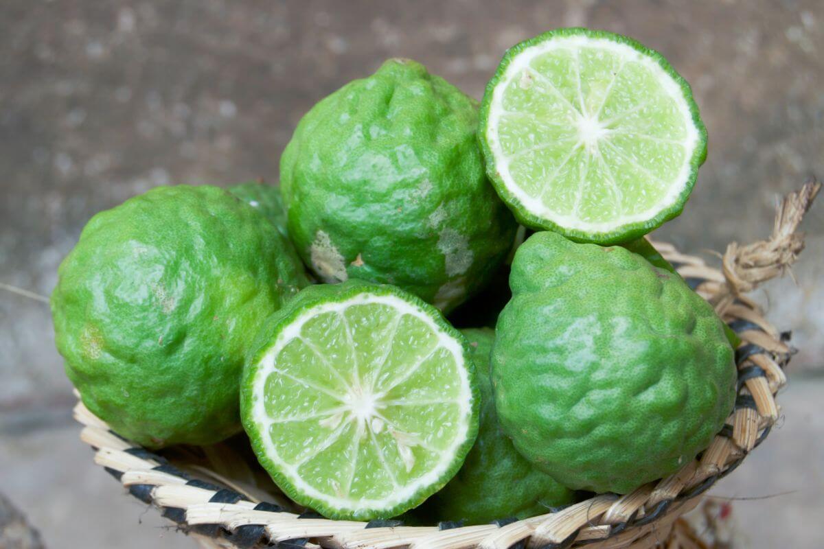 A woven basket filled with kaffir limes. Some limes are whole, showcasing their wrinkled green skin, while one lime is cut in half, revealing its juicy, segmented interior.