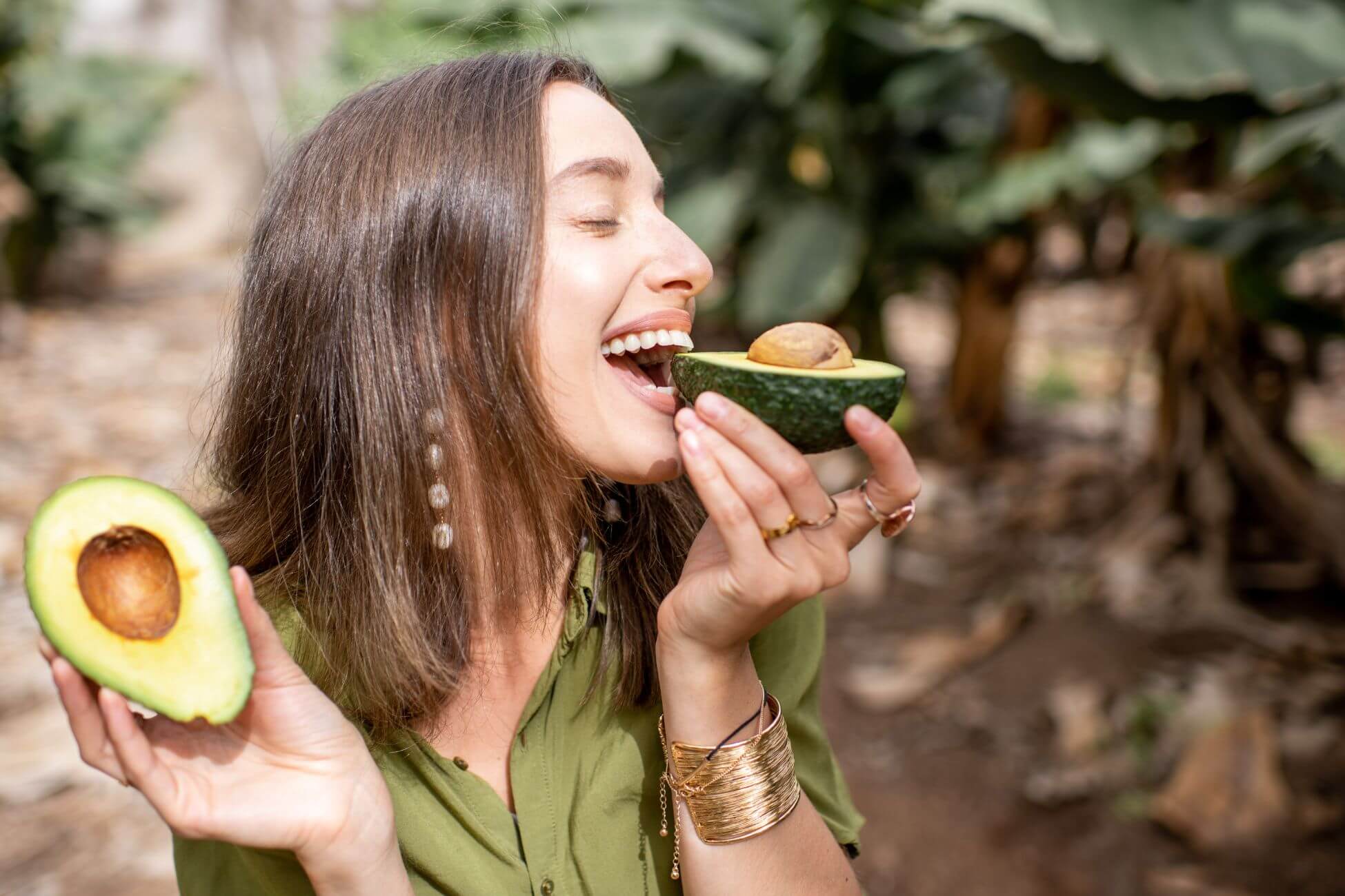 A smiling woman with shoulder-length brown hair is outdoors, holding an avocado half close to her mouth, appearing to take a bite. 
