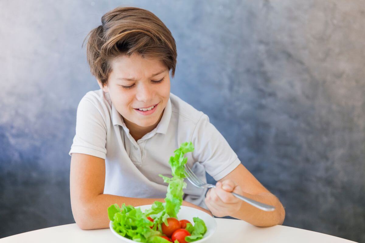 A boy in a white polo shirt sits at a table, holding a fork with a piece of lettuce, looking displeased. In front of him is a bowl of salad containing lettuce leaves and cherry tomatoes. 