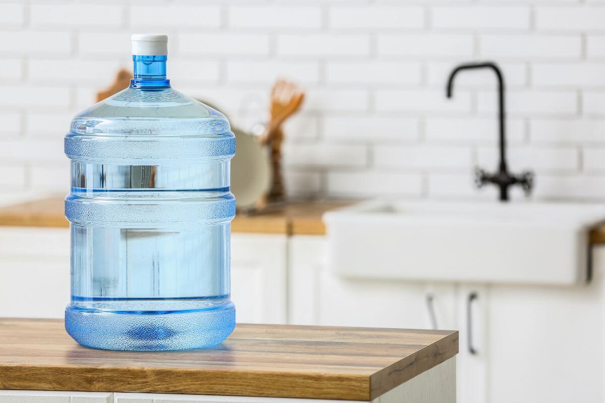 A large blue plastic distilled water jug sits on a wooden kitchen countertop. The background features a white brick wall, a white farmhouse sink, and some kitchen utensils. A black faucet is also visible in the background.