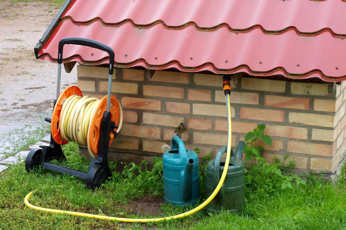 A hose connected to a tap on the side of a small brick building with a red metal roof fills two green watering cans.