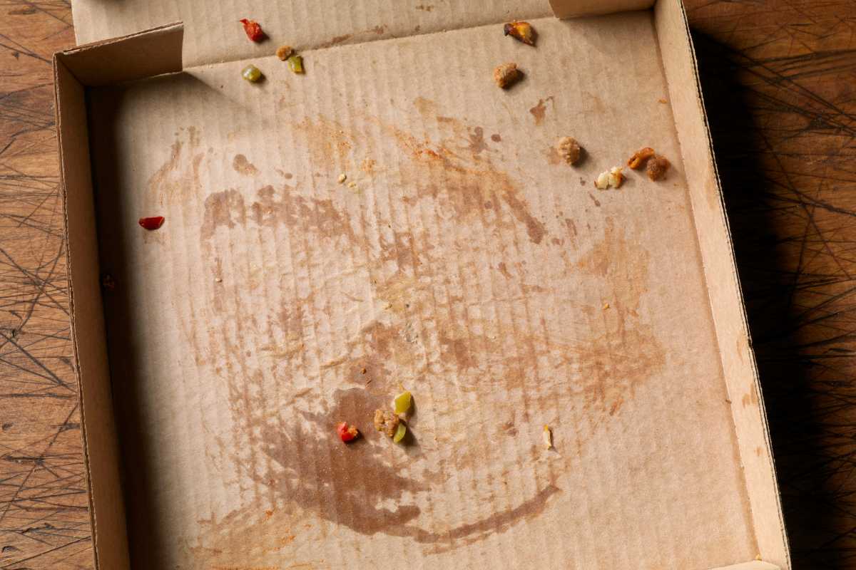 An empty pizza box with a few scattered pieces of leftover toppings such as bits of vegetables and meat, sits on a scratched wooden surface. Grease stains mark where the pizza once was. 