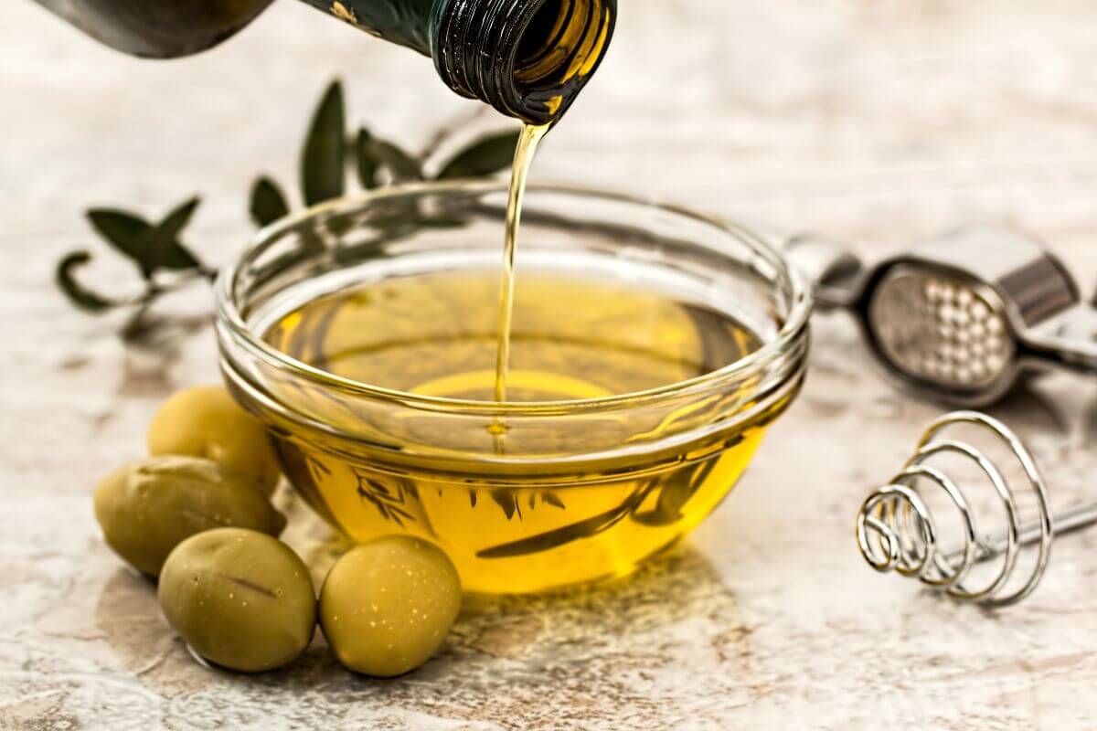 A stream of olive oil is being poured from a bottle into a clear glass bowl filled with more olive oil. Three green olives and a whisk are placed next to the bowl on the textured surface.