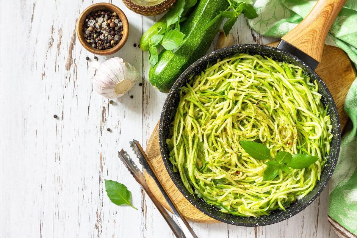 A black pan filled with cooked zucchini noodles garnished with basil leaves, placed on a wooden cutting board.