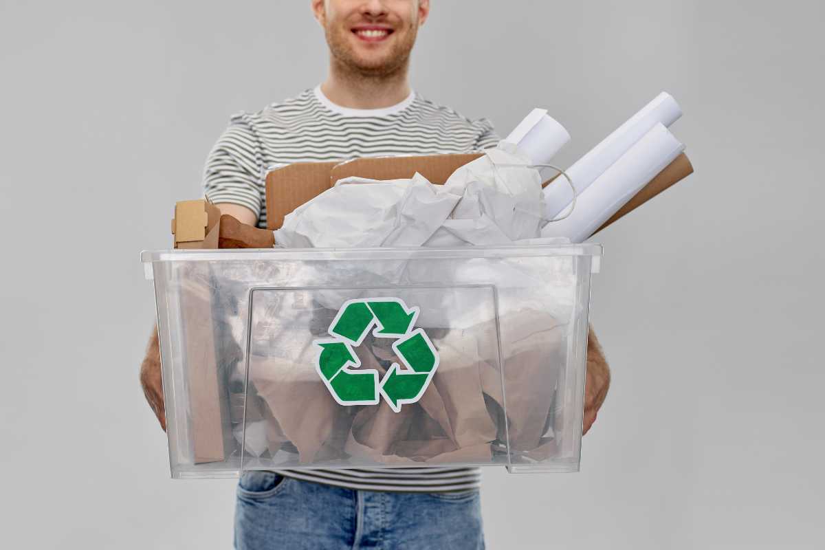 A person in a striped shirt holds a clear plastic bin filled with various recyclables, including cardboard, and papers.