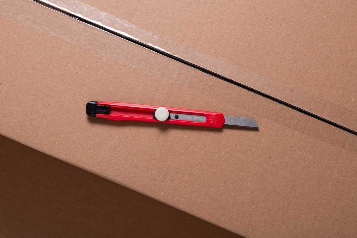 A red box cutter with a retractable blade is placed on a large cardboard box.