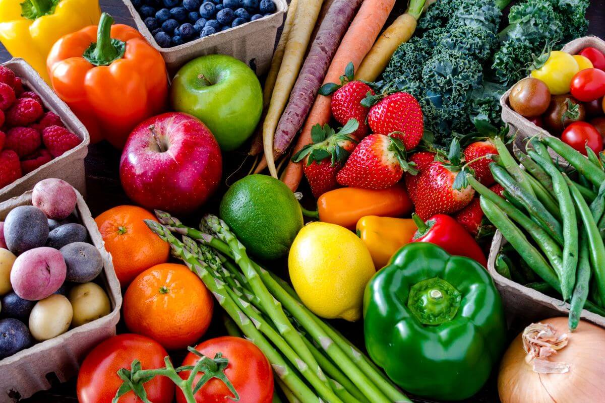 An assortment of fresh fruits and vegetables, including apples, strawberries, raspberries, blueberries, tomatoes (a classic fruit vs vegetable debate), bell peppers, carrots, asparagus, lemons, green beans, potatoes, oranges, kale, and a single onion.