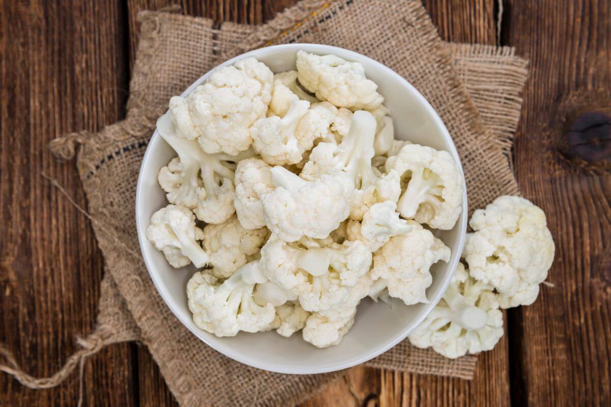 A white bowl filled with fresh cauliflower florets is placed on a piece of rustic burlap atop a wooden surface.