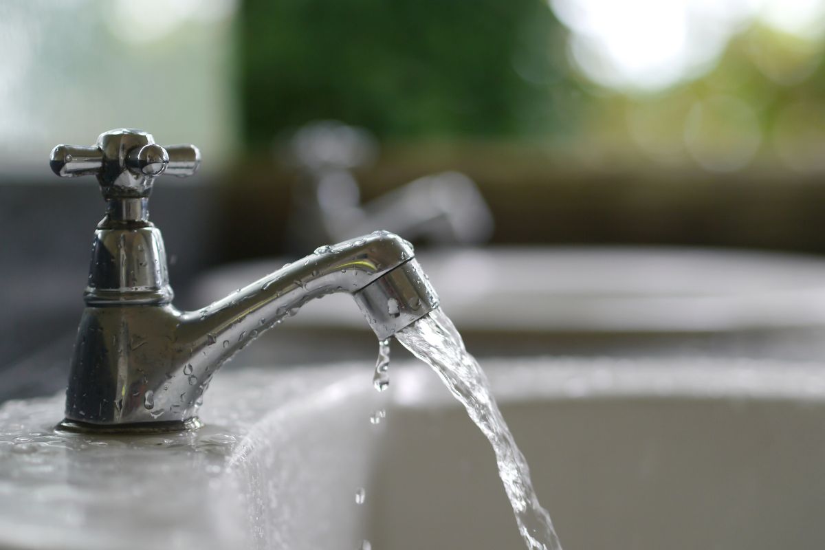 A chrome bathroom faucet with water flowing into the sink. The background is blurred but reveals another faucet.