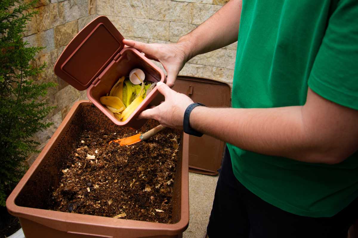 A person wearing a green shirt is adding food scraps, such as banana peels and eggshells, from a small brown container into a larger brown compost bin.