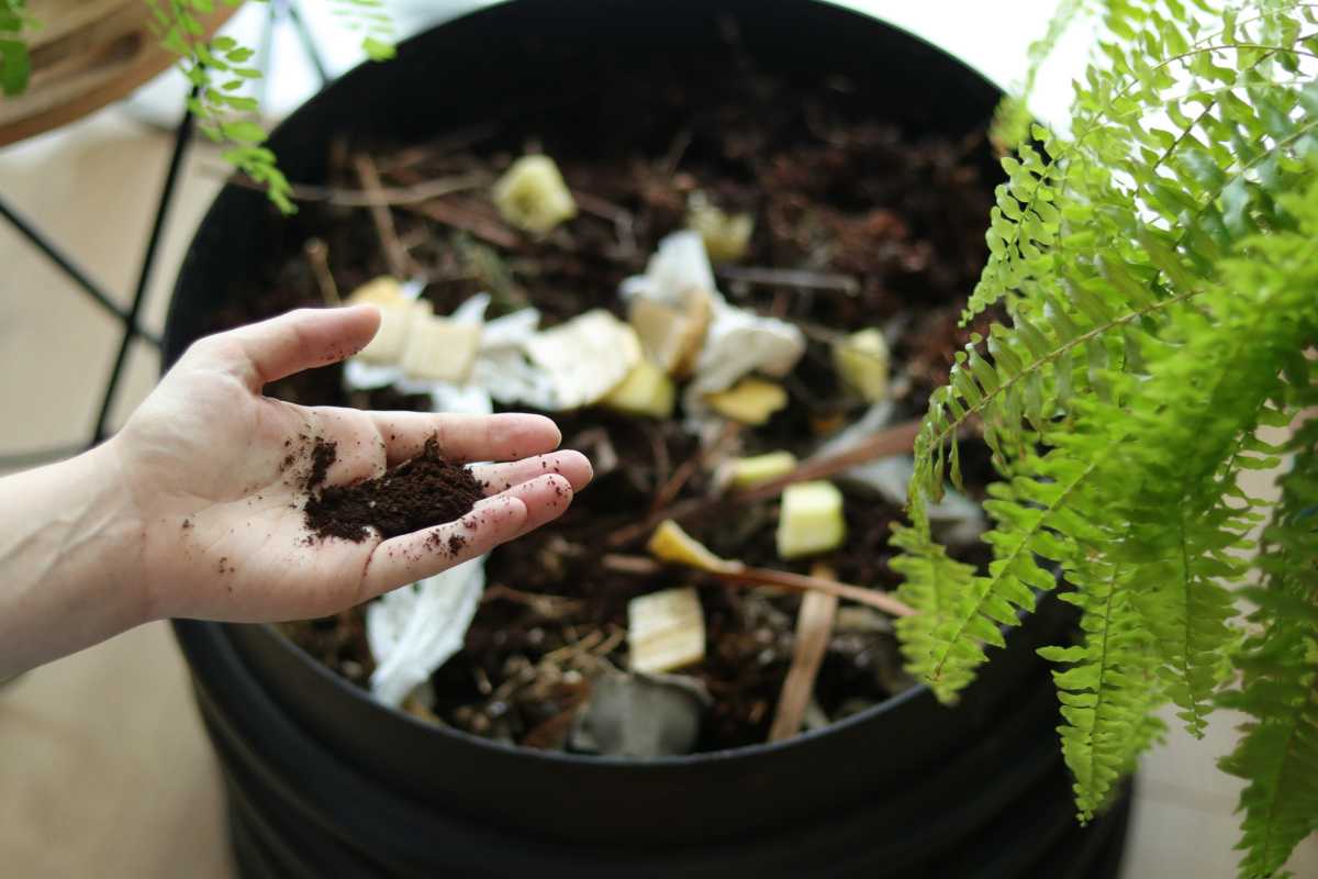 A person's hand holding soil over a large black compost bin filled with organic material, such as food scraps and paper. 