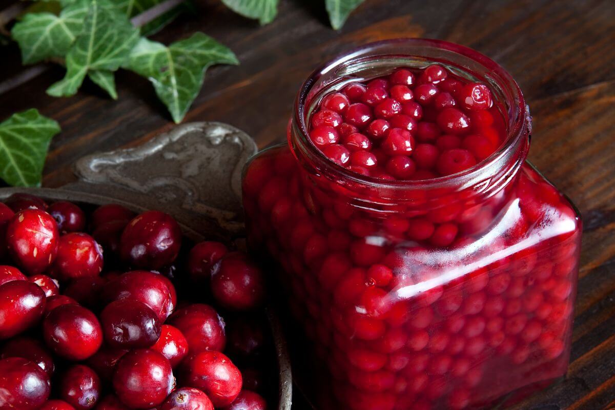 A jar filled with preserved red cranberries, sits next to a bowl of fresh cranberries on a wooden table with ivy leaves in the background.