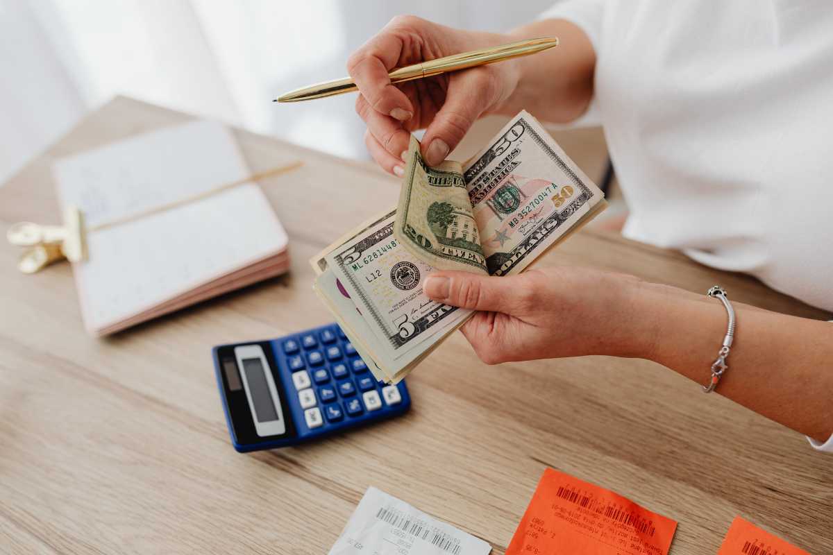 A person holding a pen and counting US dollar bills at a wooden table with a blue calculator, open notebook, and receipts nearby. 