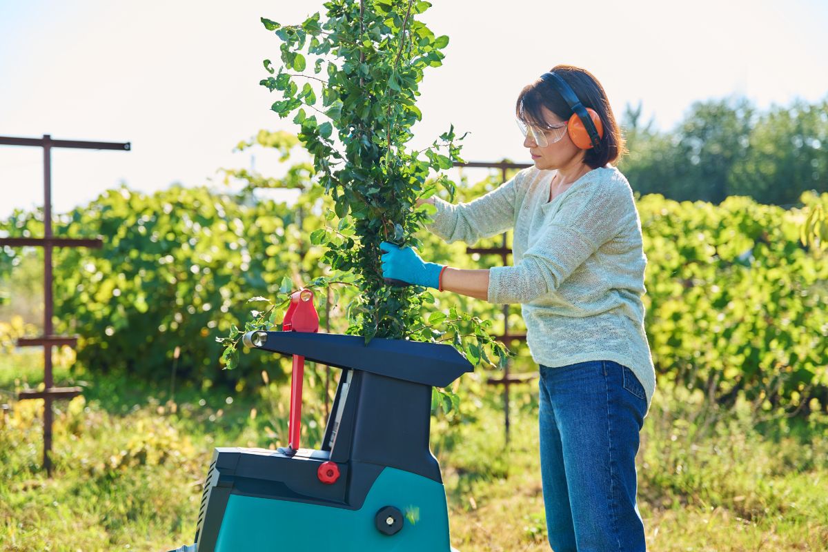 A woman wearing safety glasses, ear protection, and gloves is operating a  electric chipper shredder outdoors. 