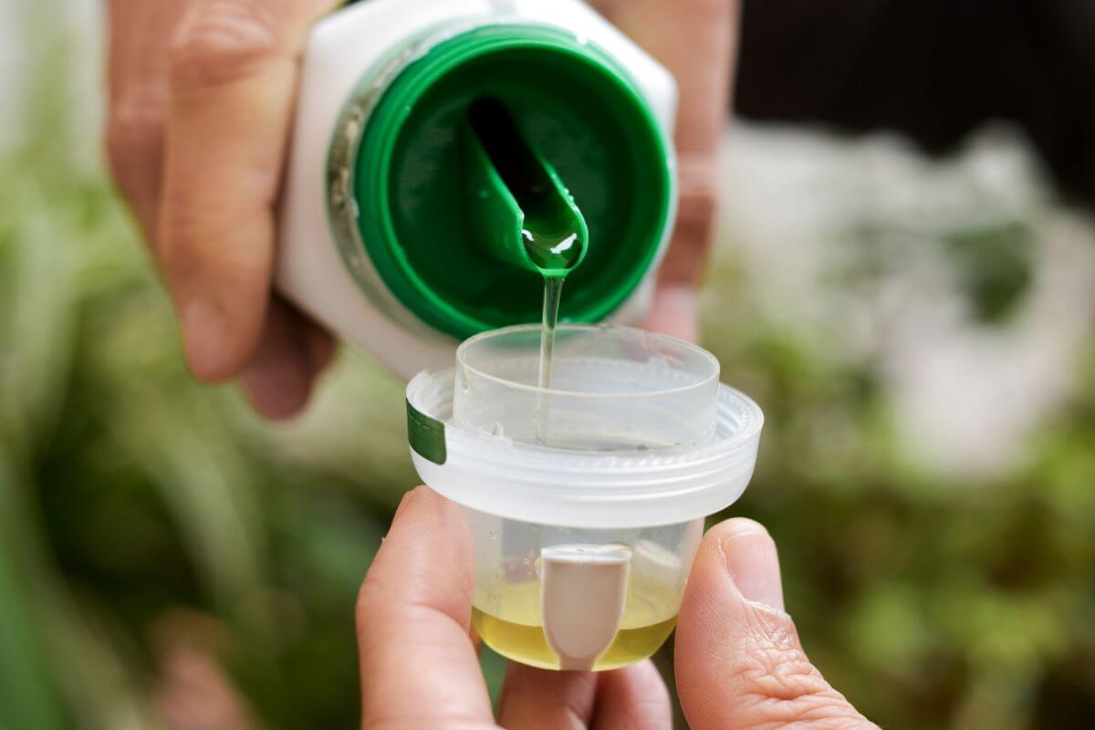 Close-up of a person pouring liquid fertilizer from a green and white bottle into a small transparent cup.