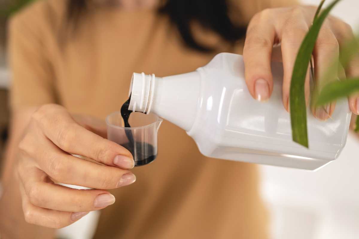 A person is pouring a dark liquid fertilizer from a white bottle into a small measuring cup, with the softly blurred background.