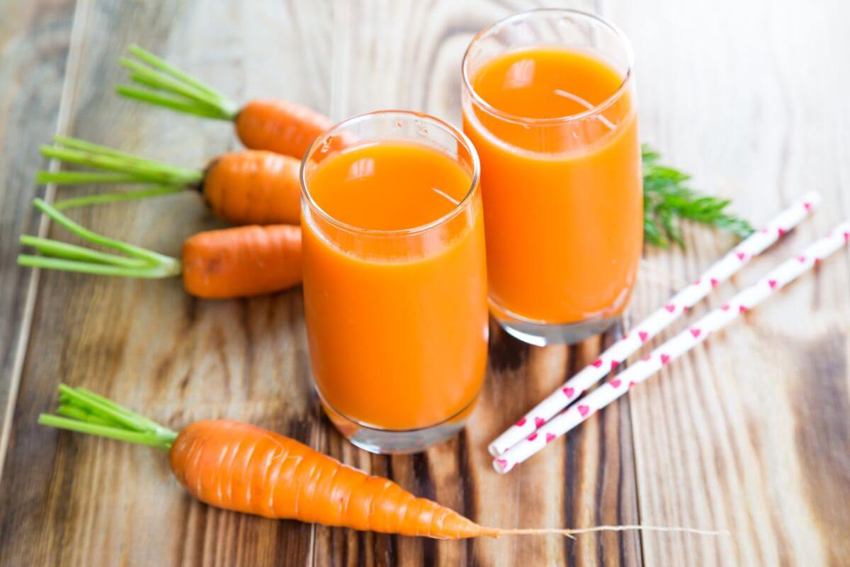 Two glasses of orange carrot juice sit on a wooden table, surrounded by several whole carrots with green tops.