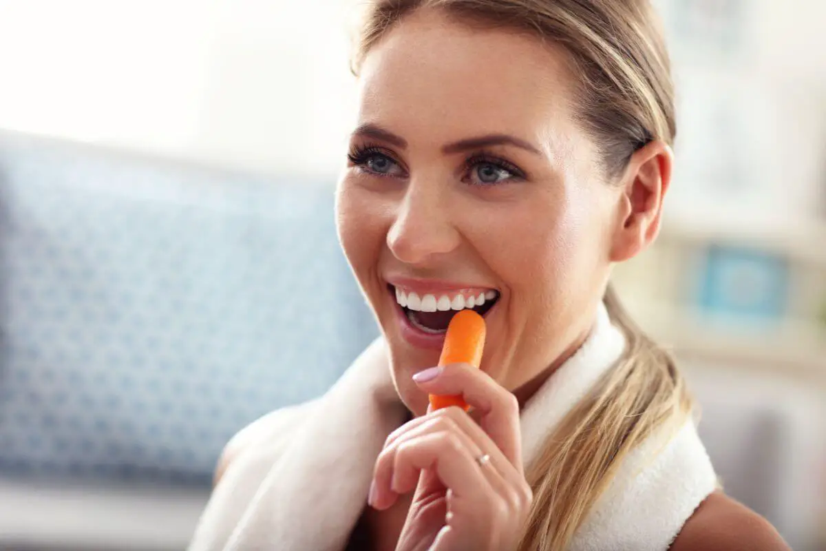 A smiling woman with long blonde hair holds a small carrot near her mouth, wearing a white towel around her neck.