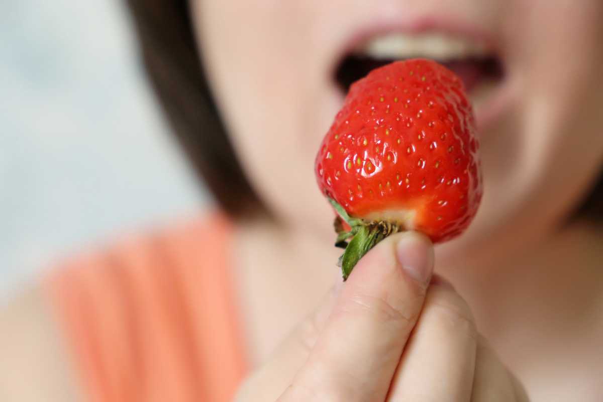 A person about to bite into a red strawberry. The strawberry is in focus, held up by the person's fingers, with the person's face slightly blurry in the background and their mouth open, 