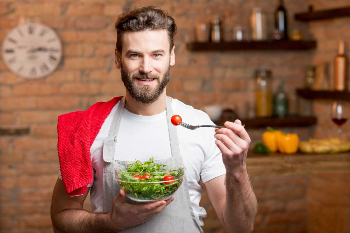 A man with a beard is standing in a kitchen, wearing a white t-shirt and a grey apron. He has a red towel over his shoulder and is holding a bowl of salad in one hand, with a fork containing a cherry tomato on the other hand.