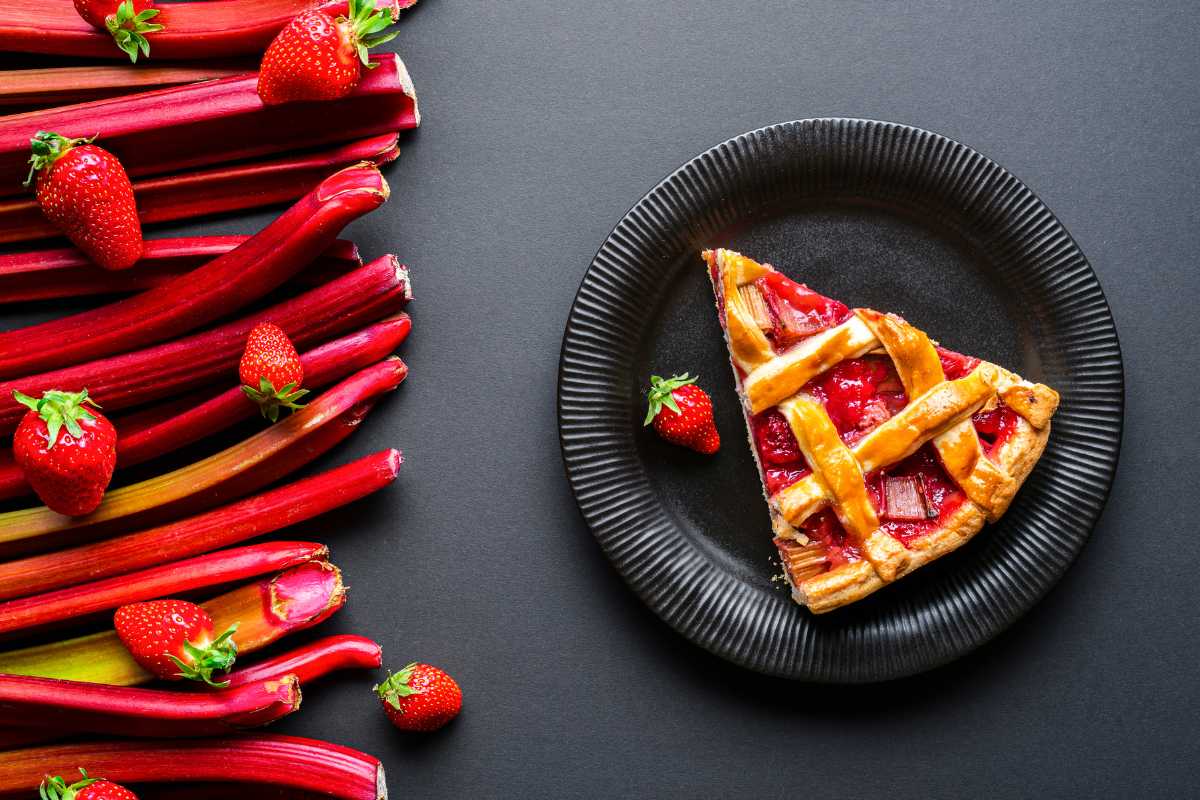 A slice of strawberry-rhubarb pie on a black plate is placed on a dark background. 