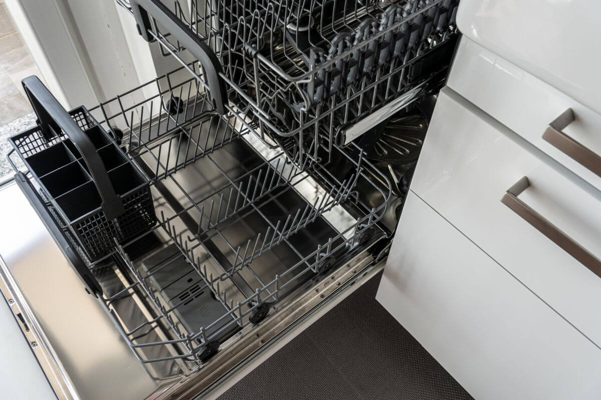 An open, empty dishwasher with two racks extended reveals its stainless steel interior and a utensil holder visibly placed on the lower rack. 