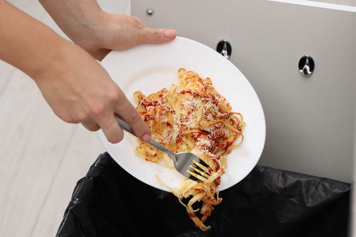 A person holding a white plate with leftover spaghetti and grated cheese is scraping the food into a black trash bin using a fork. 