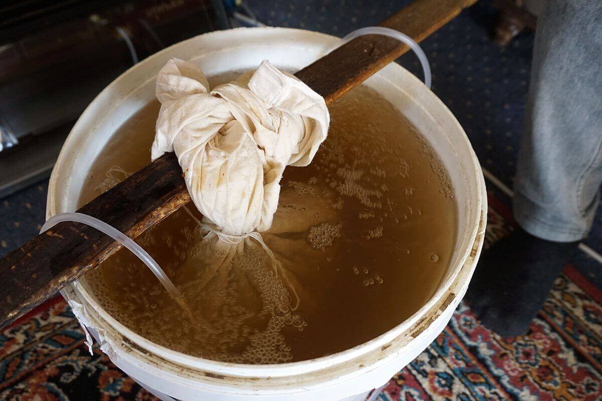 A bucket filled with worm tea showcases a wooden plank resting across its top, with a white cloth tied around the plank and extending into the liquid.