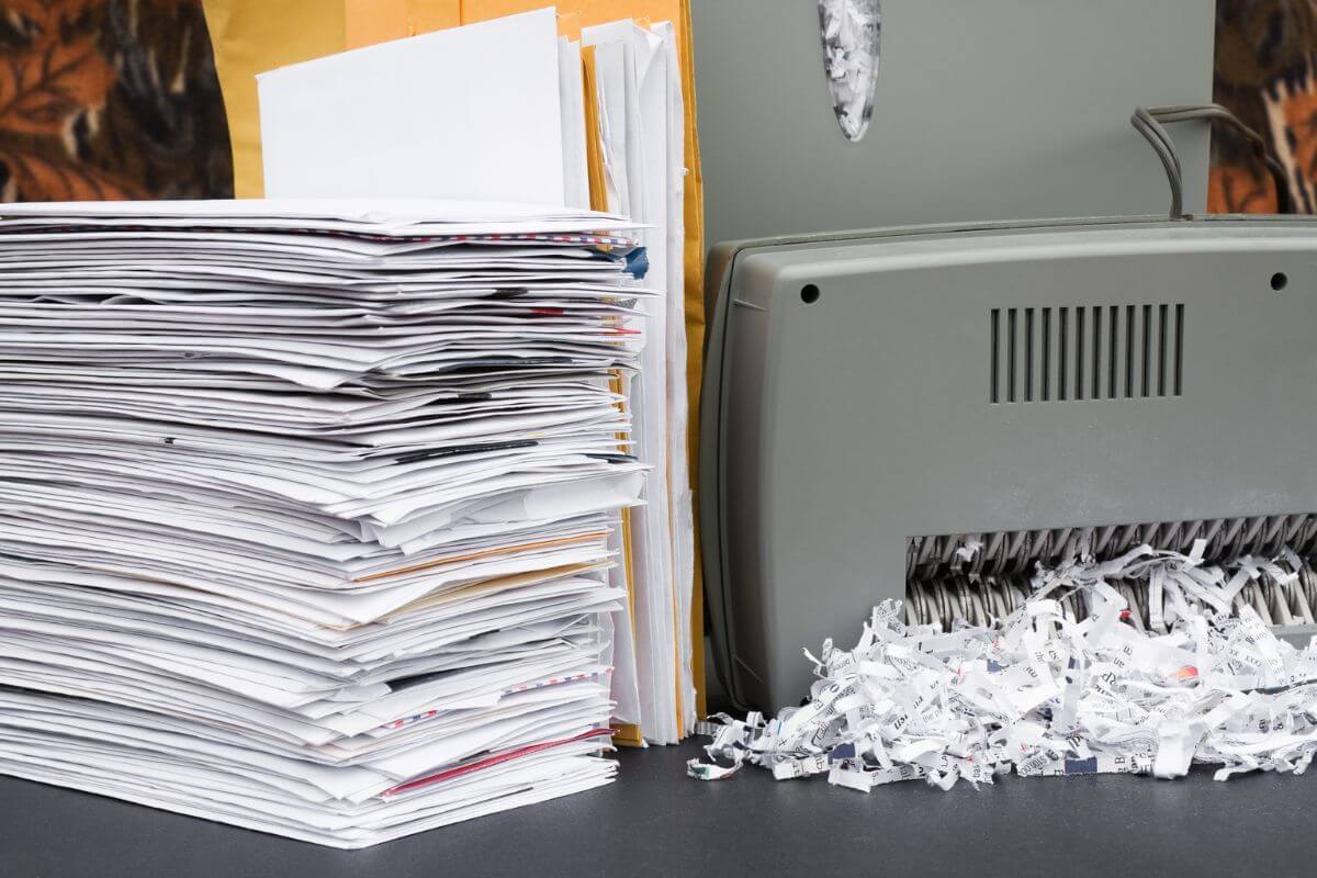 A pile of documents and envelopes sits next to a paper shredder, with shredded paper spilling out onto the surface below.