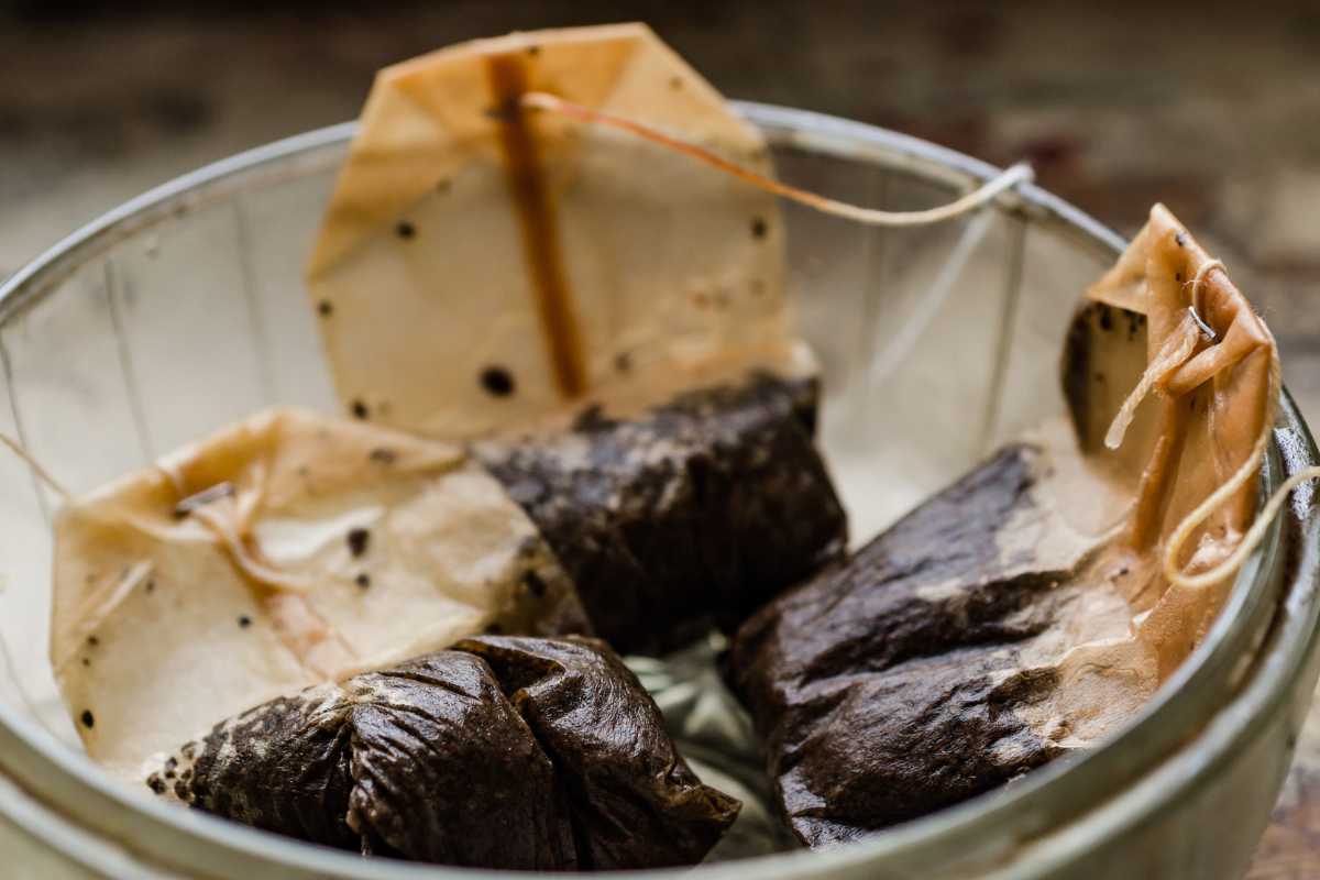 A group of used tea bags with dark stains and wrinkled paper are gathered in a glass container. The string and tags are still attached, and the tea bags appear to be damp, indicating recent use.