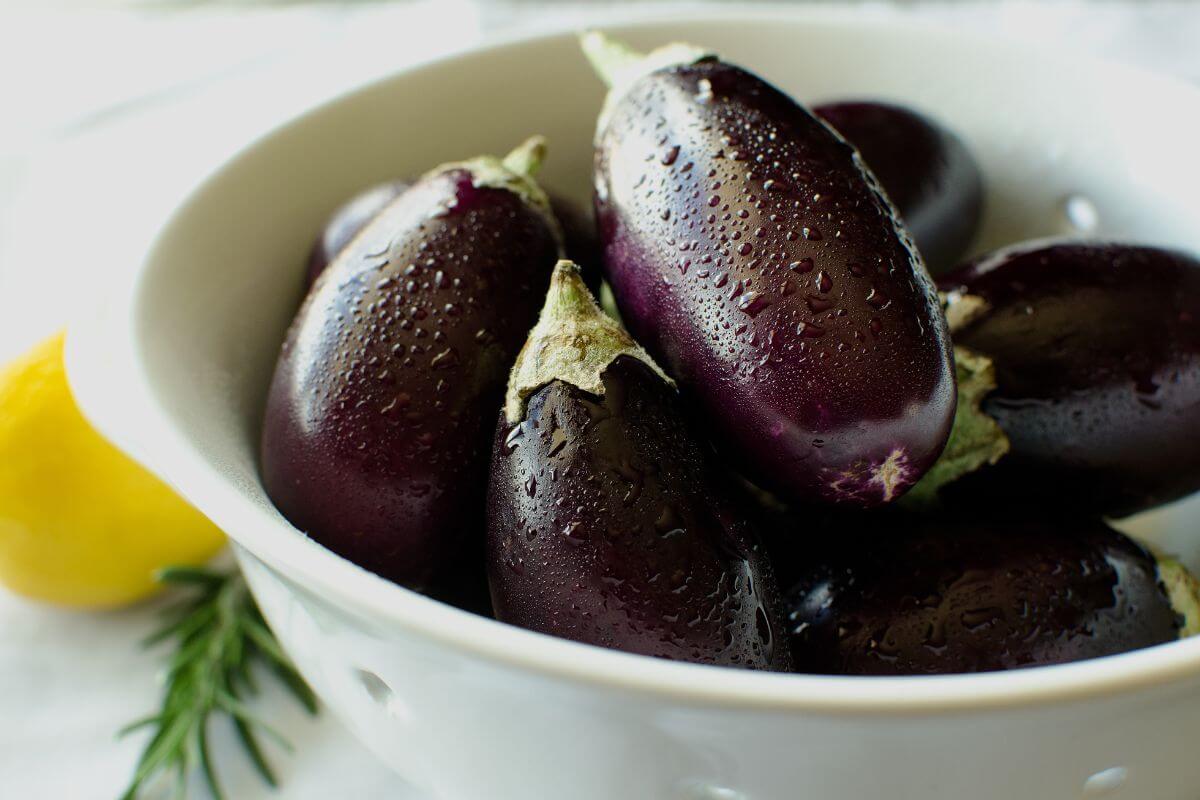 A white colander holds fresh Indian eggplants, their dark purple skins gleaming with water droplets, contrasted by vibrant green stems. 
