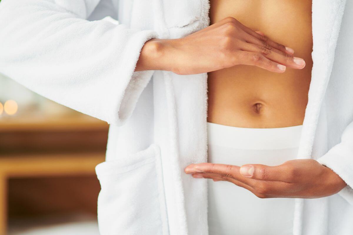 A person wearing a white bathrobe places their hands in a circular shape in front of their bare midriff, highlighting their abdomen.