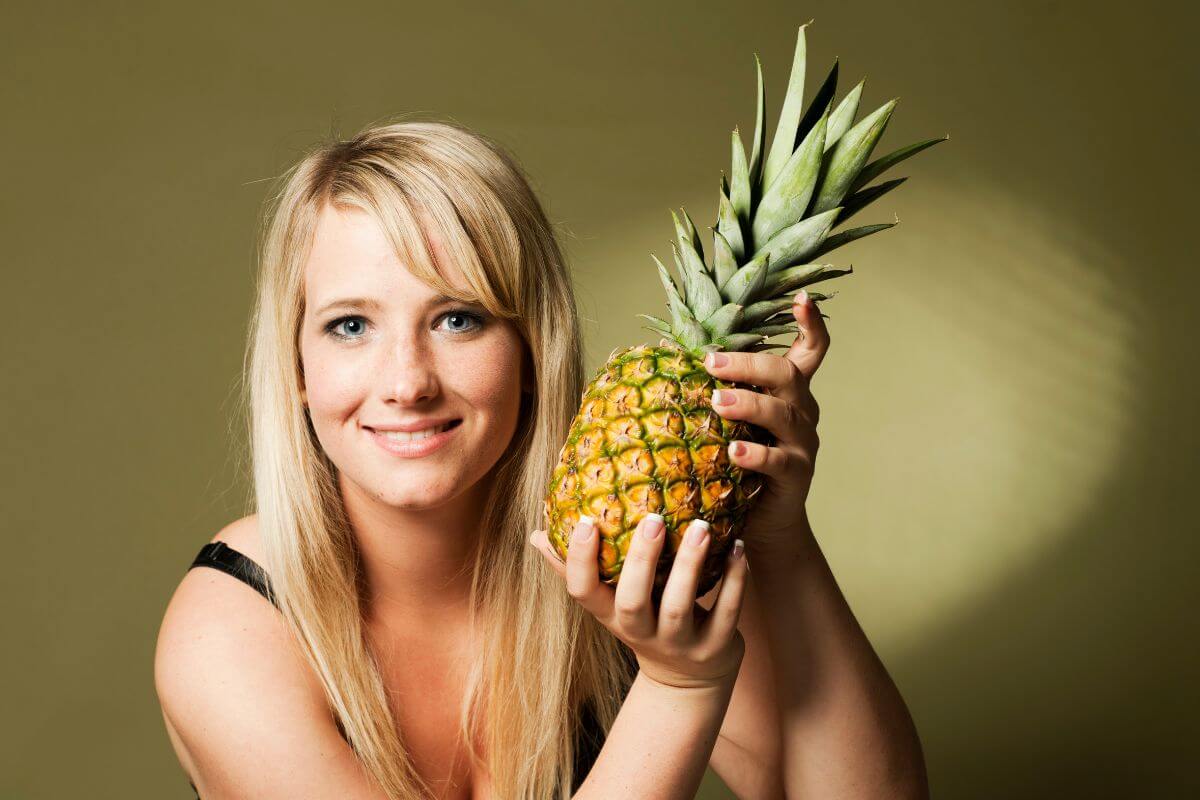 A smiling woman with long blonde hair holds a ripe pineapple close to her face.