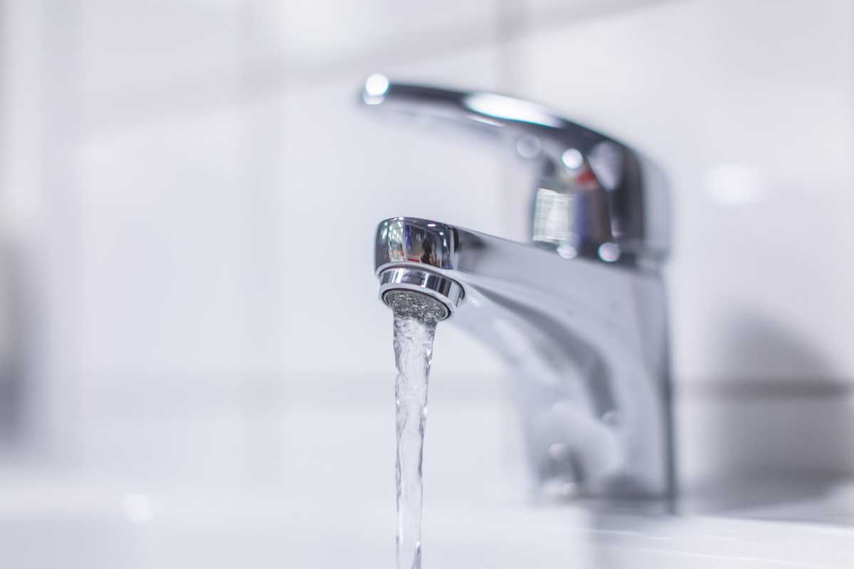 A modern, chrome-finished faucet with water flowing out, set against a blurred white tiled backround.